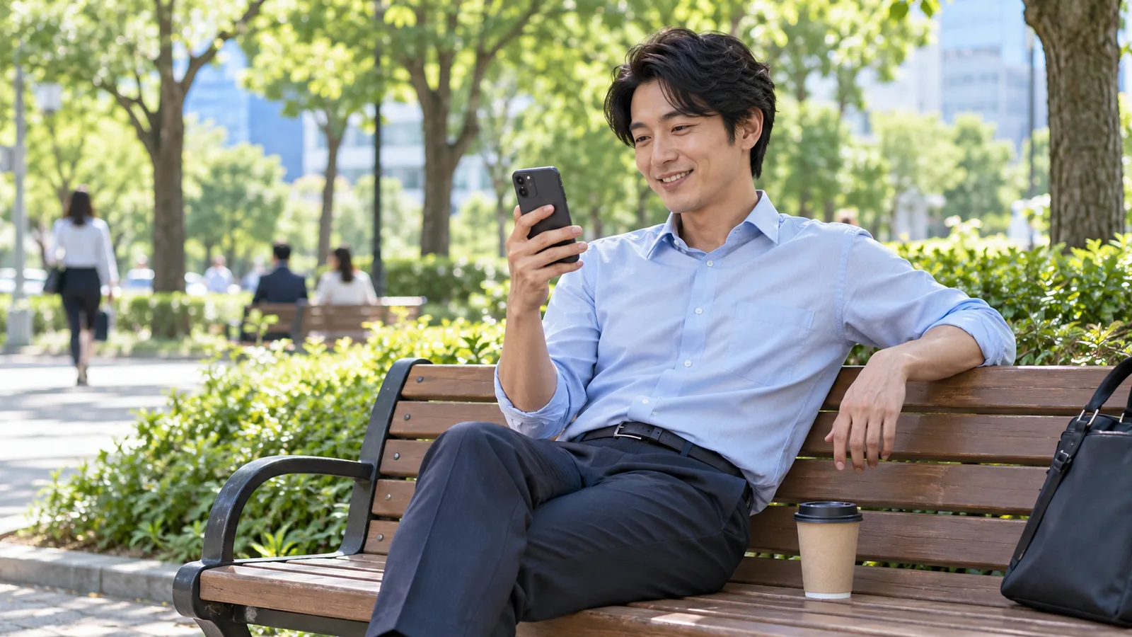 緑の多いオフィス街の公園のベンチで、男性が笑顔でスマートフォンの動画を見ている様子