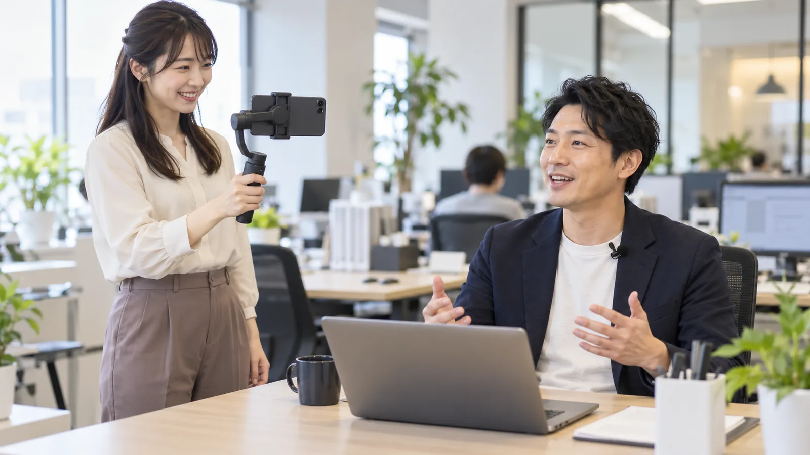 明るいオフィスで、女性がジンバルに付けたスマートフォンで同僚の話す様子を撮影している様子